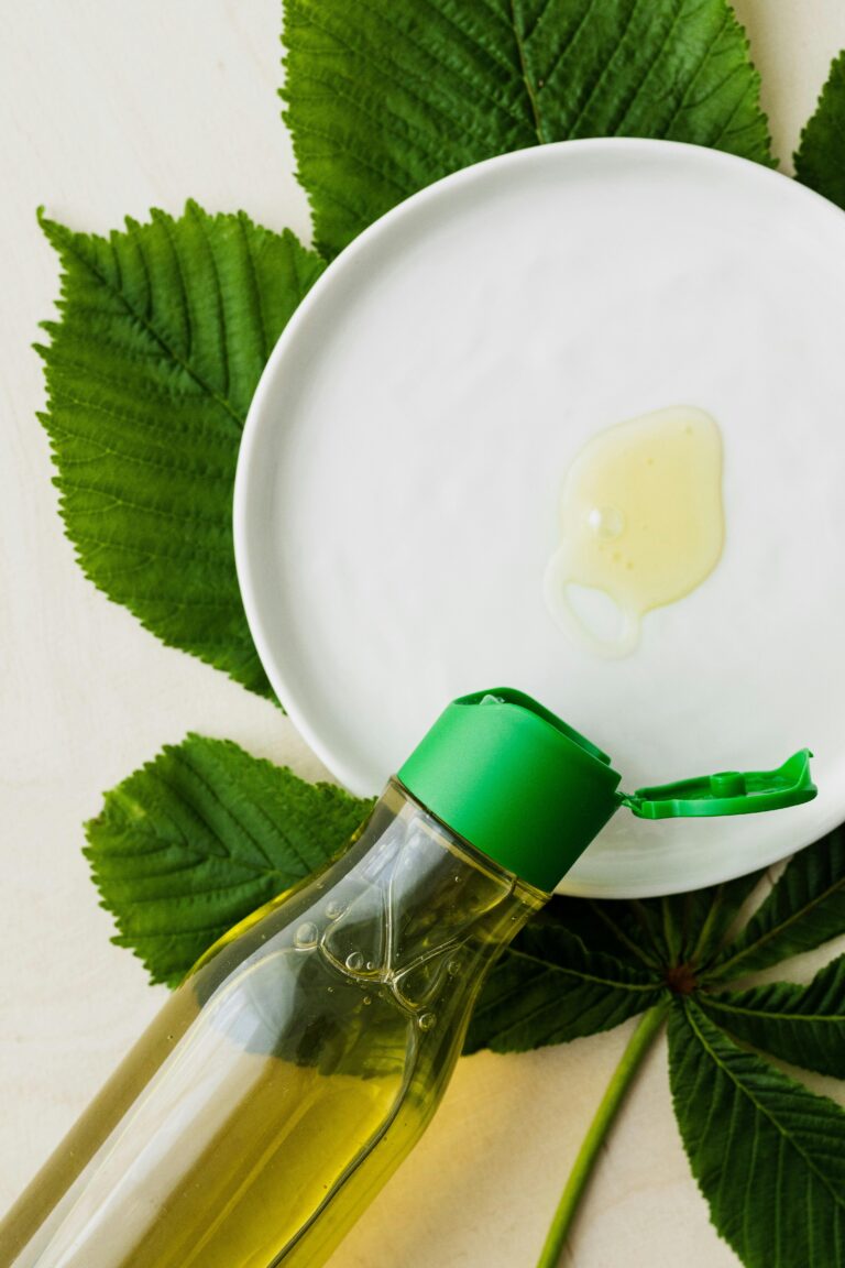 Nosotros Top view of open bottle of cosmetic oil placed flat on table near white plate with small amount of oil on it on big fresh green leaf